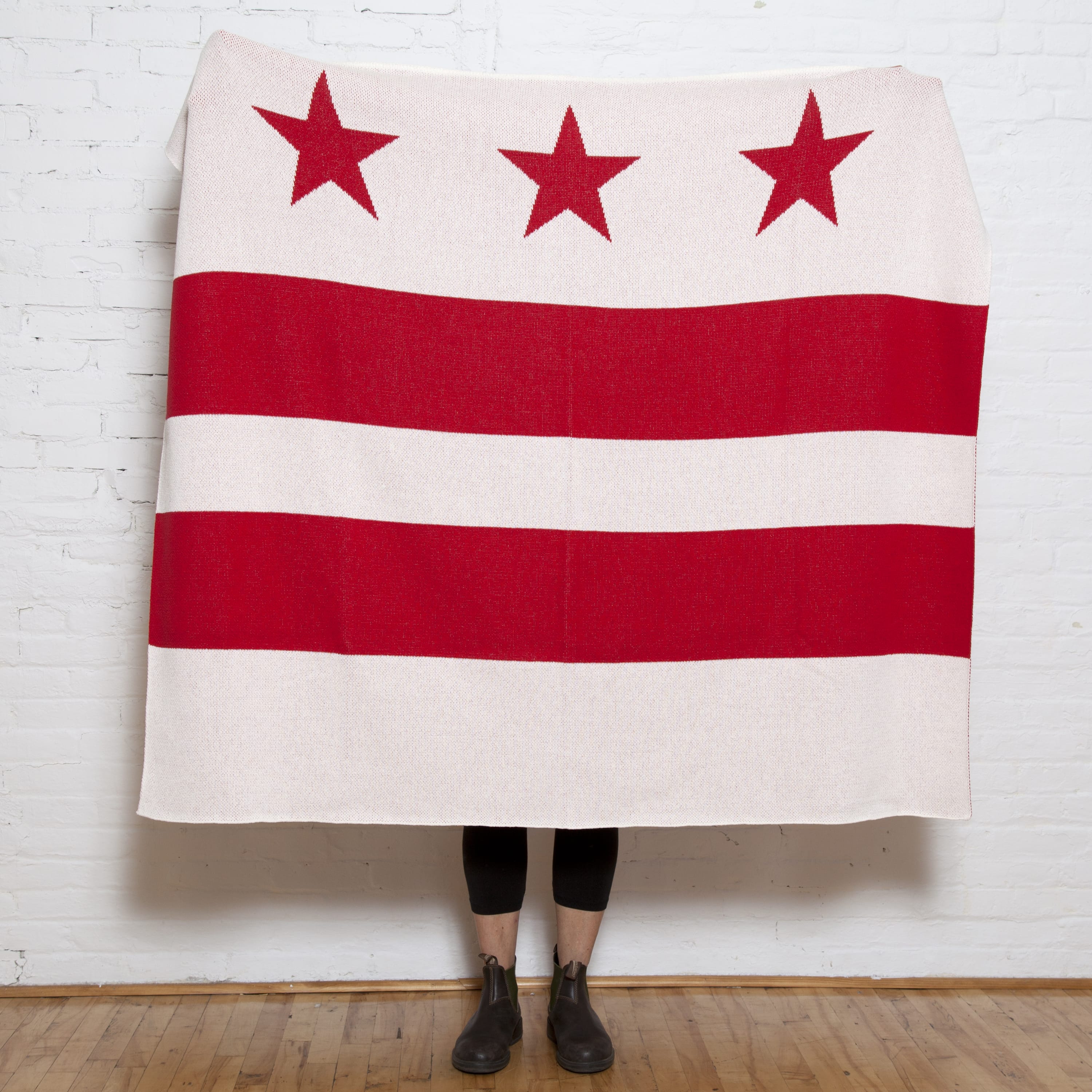 A person stands behind the in2green Hometown Pride Flag - Cotton Sustainable Knit Blanket, featuring the Washington, D.C. flag design with three red stars and two red bars on a white background; only their lower legs and feet are visible. This custom blanket is proudly made in the USA from recycled cotton.