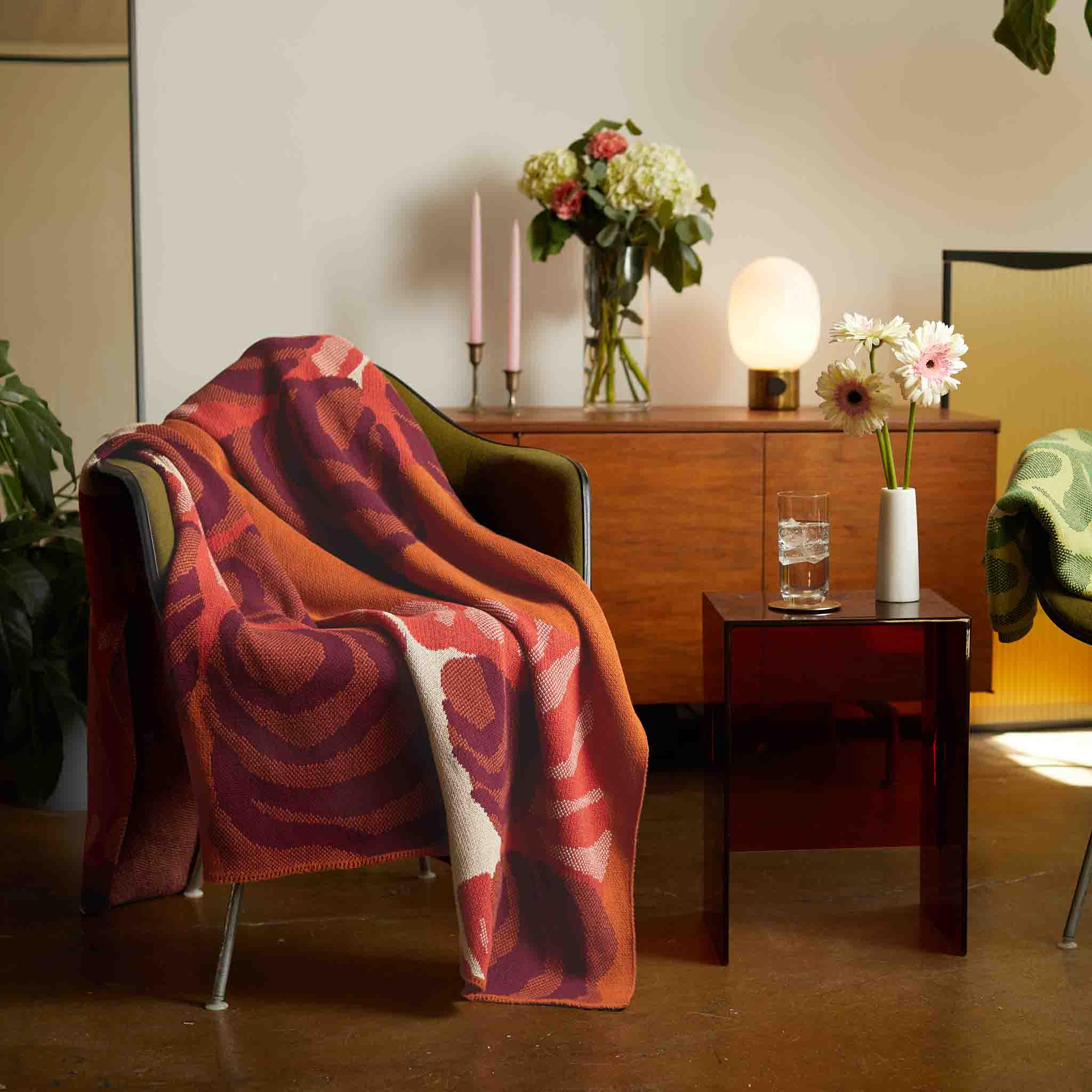 A chair is draped with the Floréal 7 Throw Blanket by E.A. Seguy from In2Green, featuring red and pink patterns; beside it, a small table holds a vase of flowers and a glass of water, with a sideboard adorned with flowers and candles in the background.