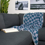 The in2green Gingham Plaid Throw Blanket in blue and white is draped over a dark gray sofa beside an open book, with plant and beach photos hanging on the wall in the background.