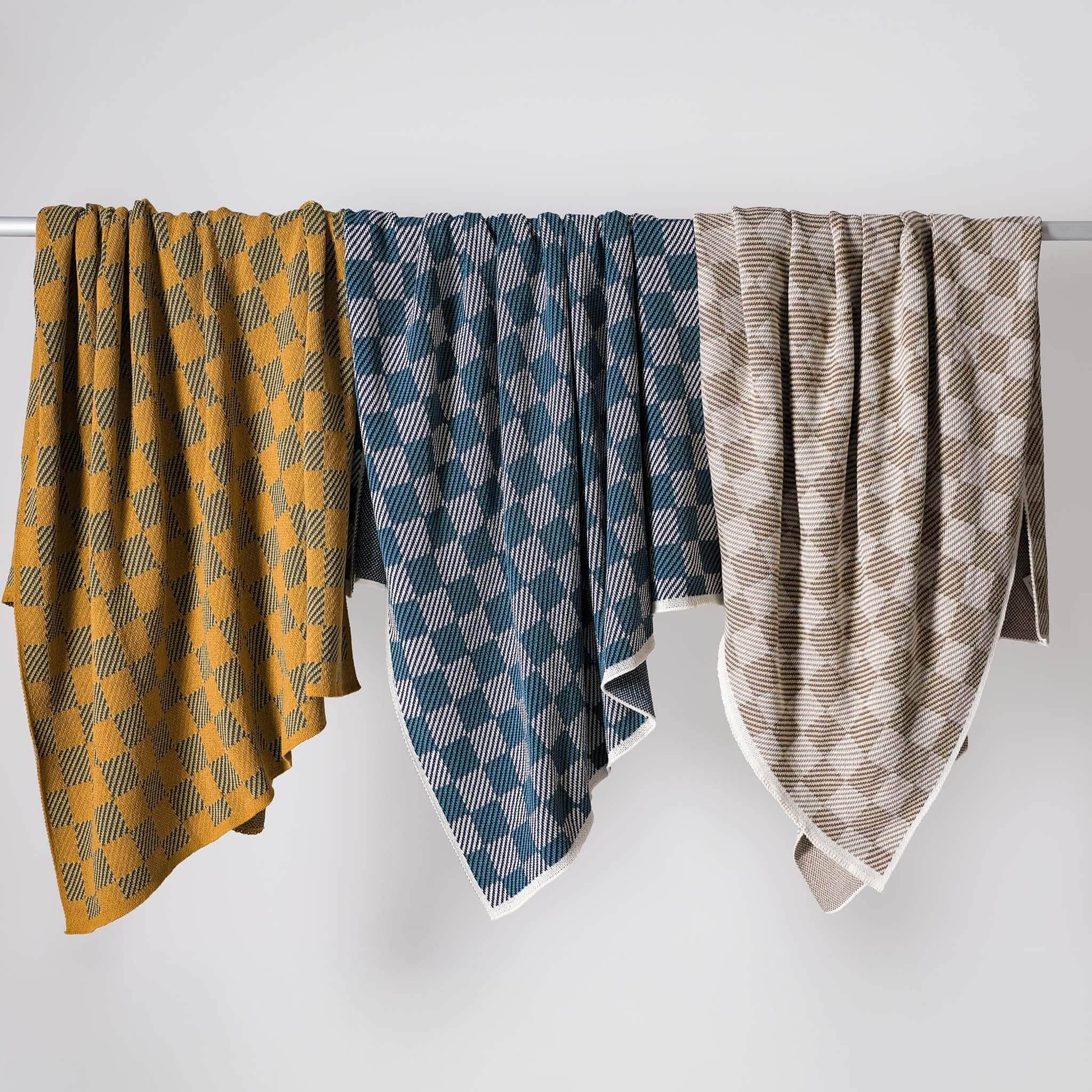 Three patterned blankets—including the in2green Gingham Plaid Throw Blanket—hang side by side over a horizontal rod, displayed against a plain white background.