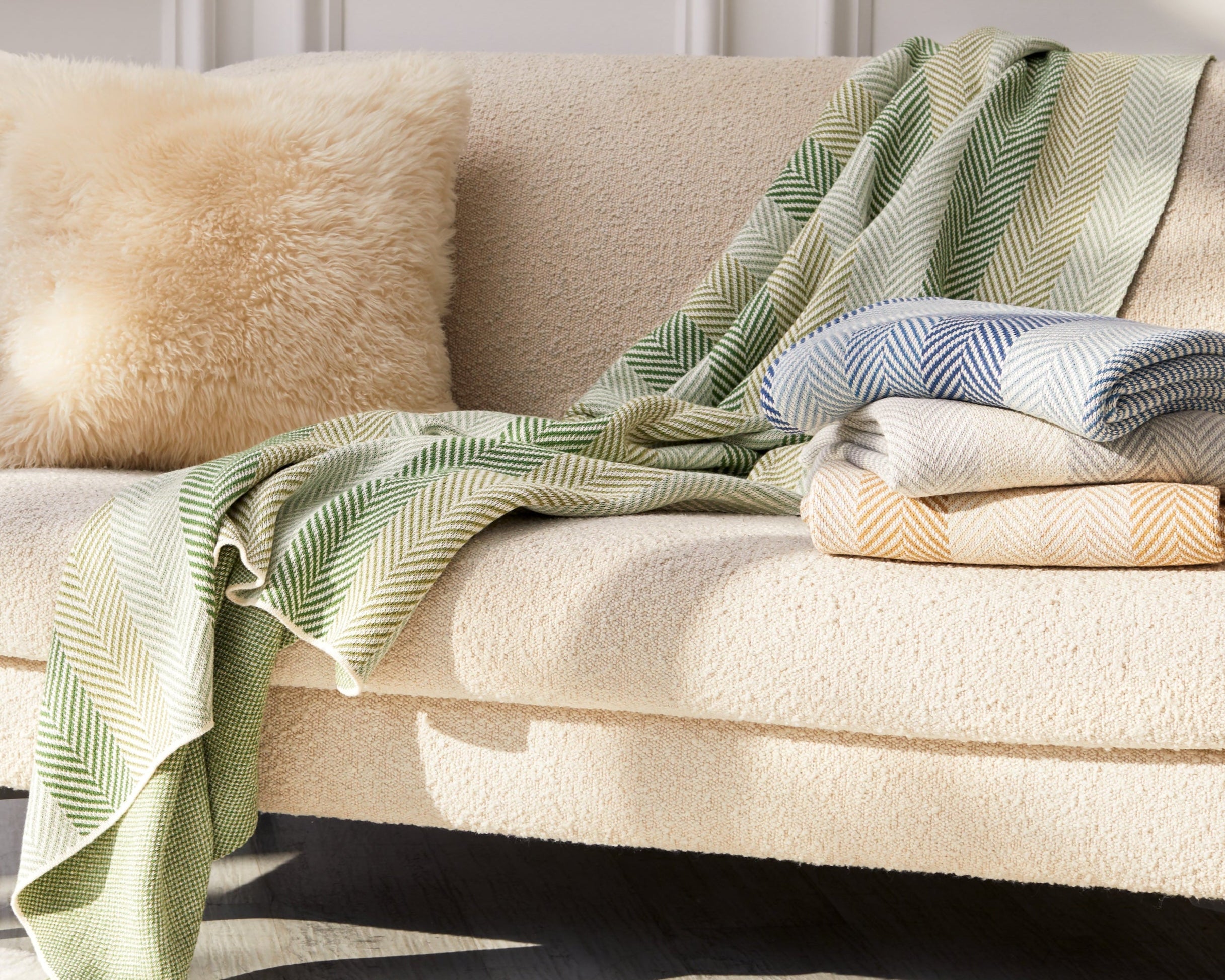 A cream-colored sofa with a fuzzy pillow, the Herringbone Hues Throw Blanket made from recycled yarns in a green pattern draped over the side, and two folded blue and beige blankets on the seat, set in a bright, elegant room.