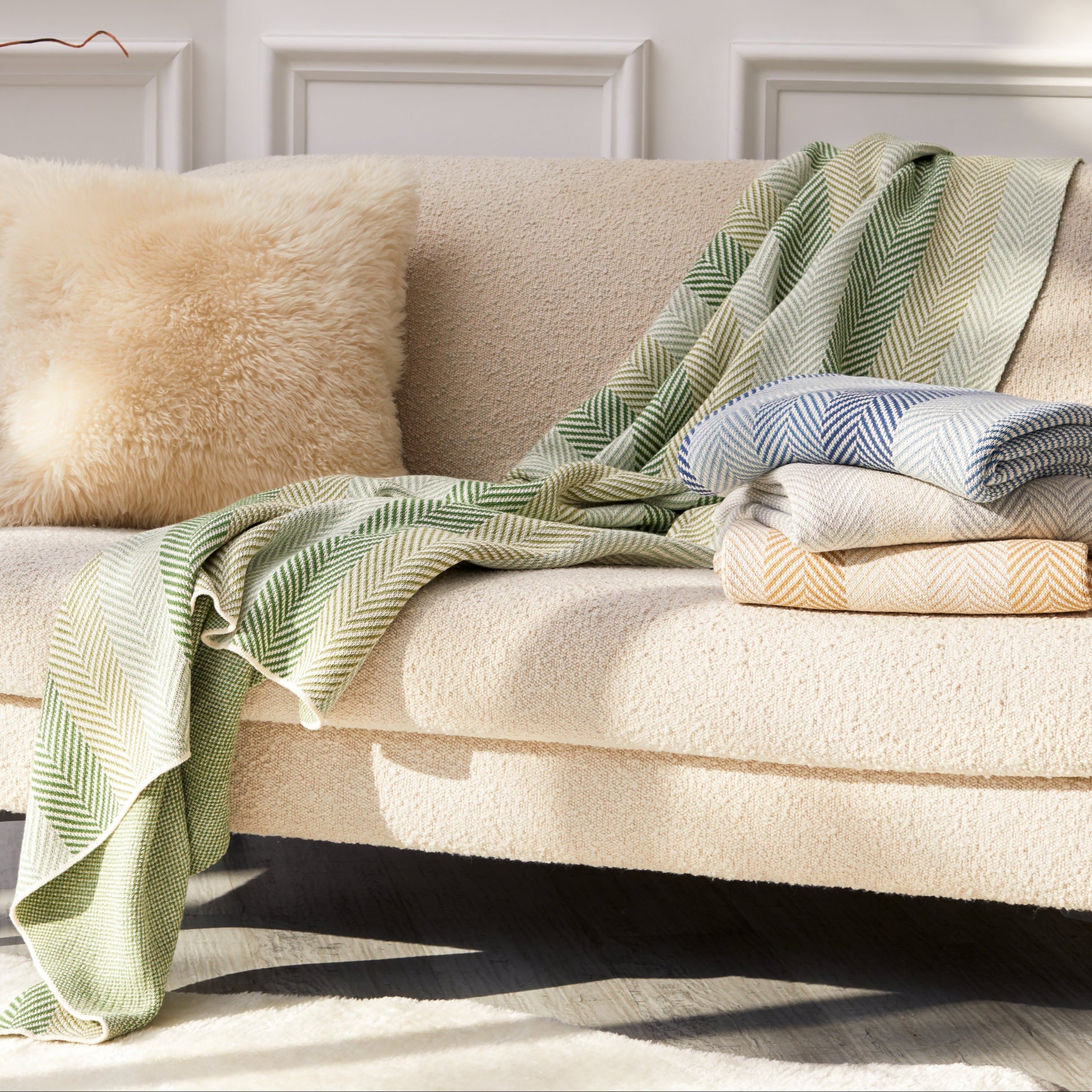 A cream-colored sofa with a fuzzy pillow, the Herringbone Hues Throw Blanket made from recycled yarns in a green pattern draped over the side, and two folded blue and beige blankets on the seat, set in a bright, elegant room.
