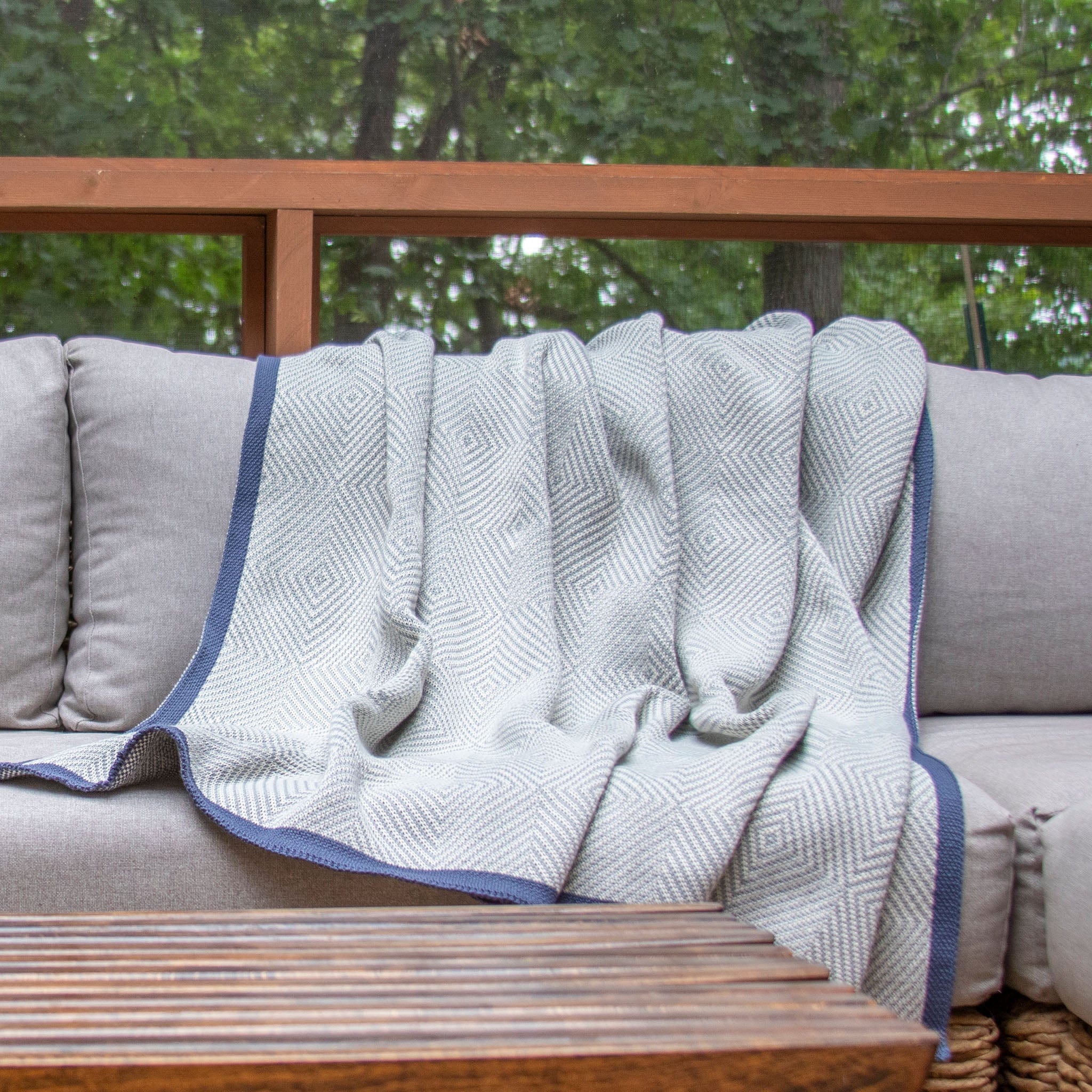 The in2green Poly Woven Square Throw Blanket, made from recycled plastic bottles and featuring a light pattern with a dark border, is draped over a gray outdoor sofa beside a wooden table, creating a cozy space with trees in the background.