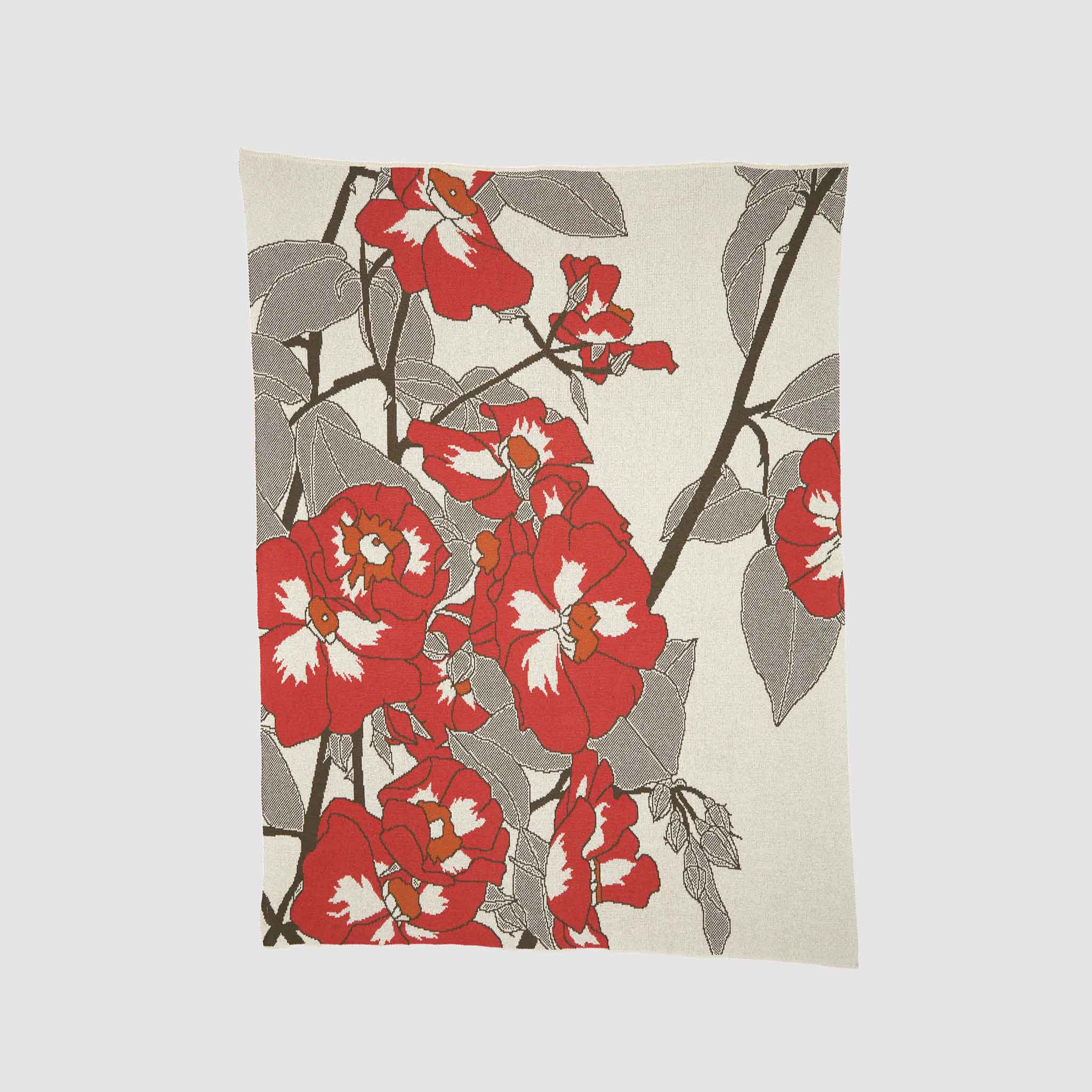 The Prairie Rose Throw Blanket by Hannah Borger Overbeck for in2green is a rectangular recycled cotton blanket with a prairie rose design of red and white flowers, brown branches, and gray leaves on a light background.