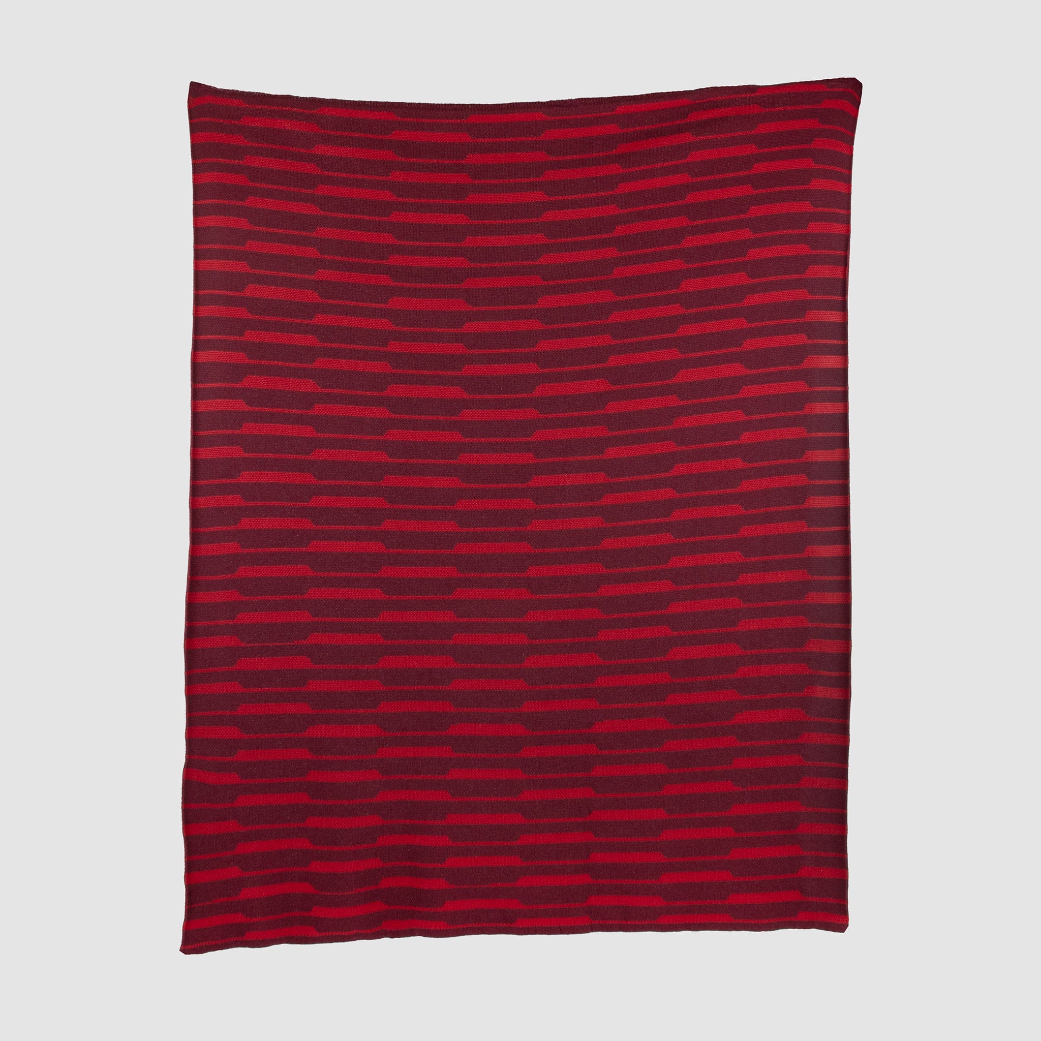 The Riata Throw Blanket by Petra Kaksonen is a rectangular red piece inspired by Scandinavian heritage, showcasing horizontal stripes in alternating dark and bright red on a plain background.