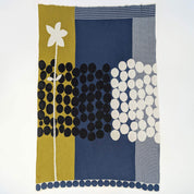 The “Rock Rose” throw by Susy Pilgrim Waters for In2Green features a modern design with a large white flower, black and white polka dots, and sections of olive green, navy blue, and patterned backgrounds.