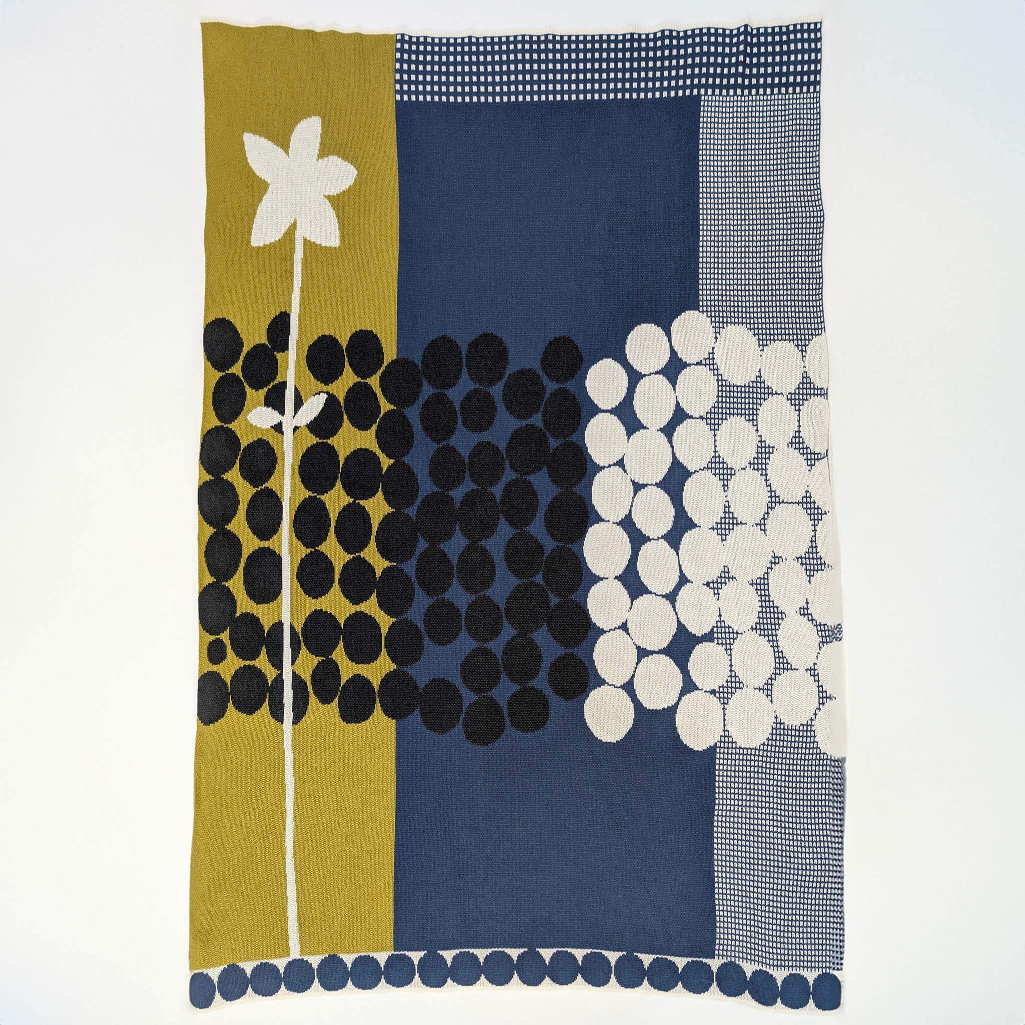 The “Rock Rose” throw by Susy Pilgrim Waters for In2Green features a modern design with a large white flower, black and white polka dots, and sections of olive green, navy blue, and patterned backgrounds.