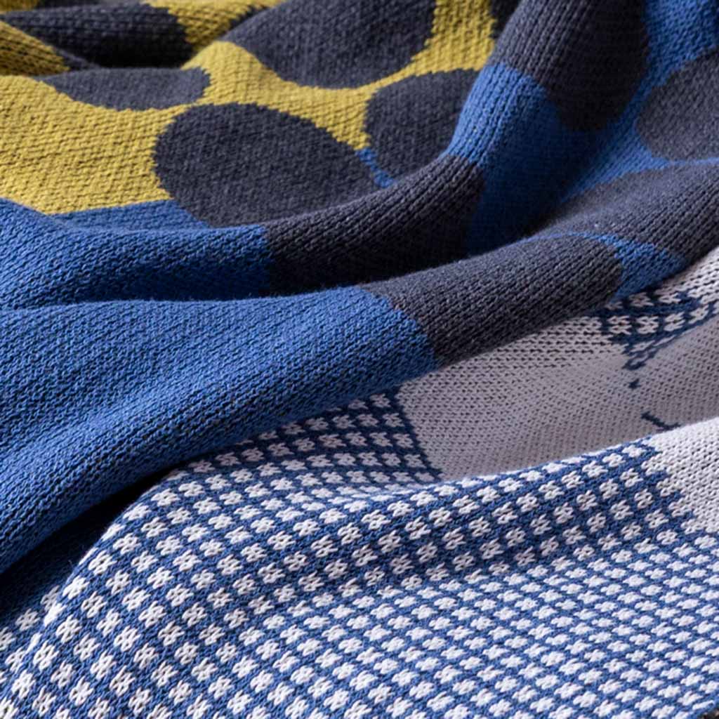 A close-up of the Cotton Sustainable Knit Floral | Graphic Throw Blanket - Rock Rose by Susy Pilgrim Waters, from In2Green, showcases modern blue, yellow, black, and white geometric and polka dot patterns with a hand-crafted feel.
