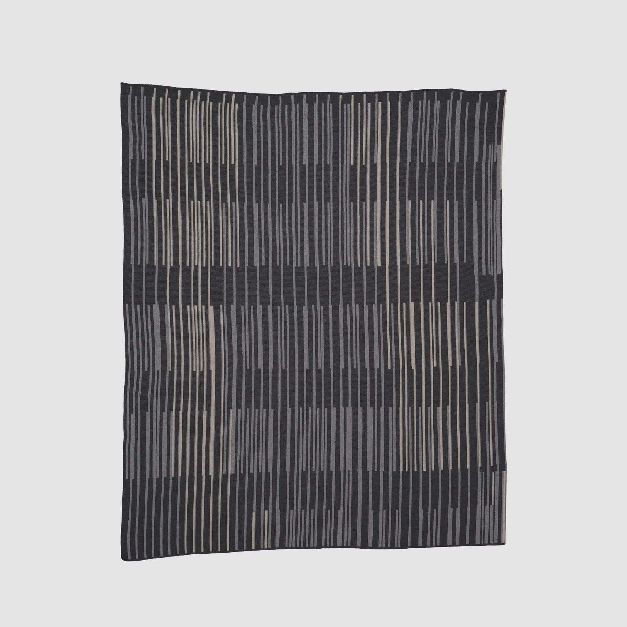The Washed Throw Blanket by Jill Malek for In2Green features a black background with vertical beige and gray stripes in an irregular, nature-inspired pattern, reflecting Malek's organic design style.