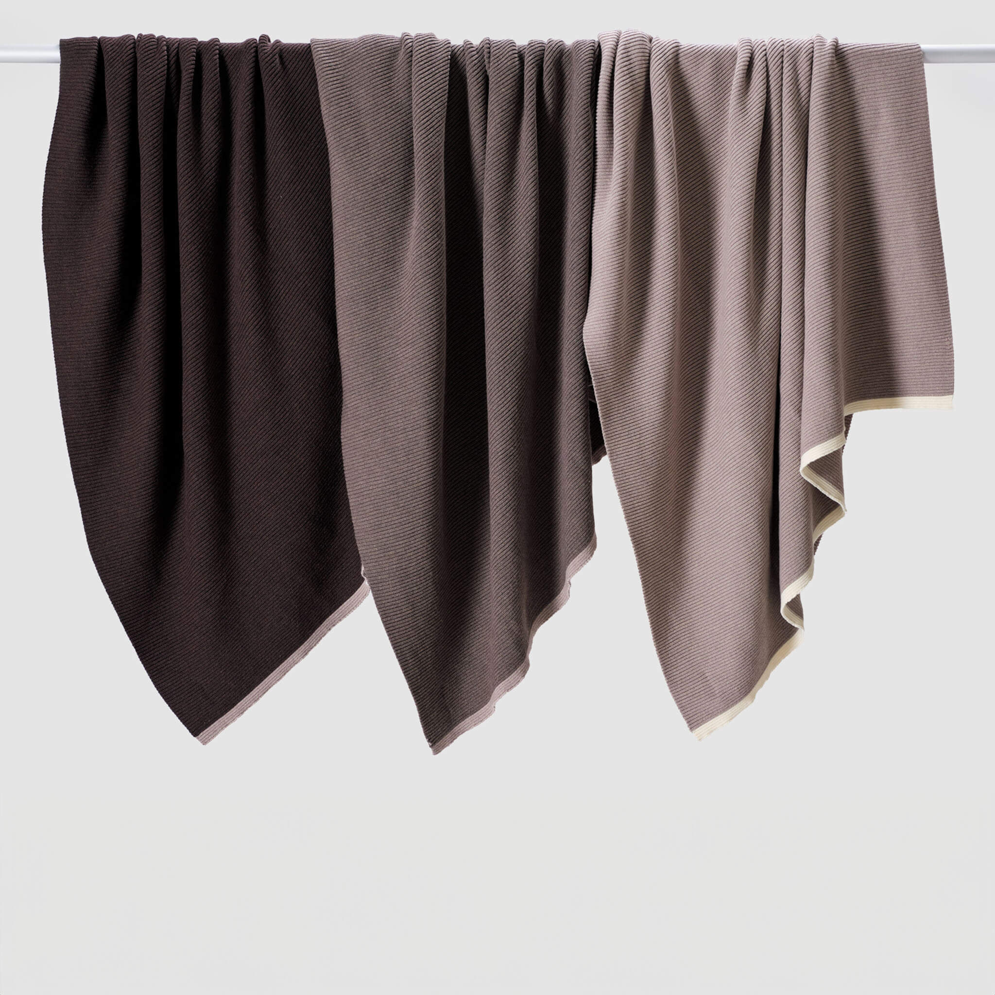 Three textured blankets hang side by side on a rod, including the in2green 100% Merino Wool Links Throw Blanket in minimalist shades of brown and beige, displayed against a plain background.