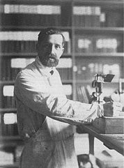 A man in a lab coat stands beside a microscope, with bookshelves behind. The Art Nouveau decor, featuring the Floréal 7 Throw Blanket by E.A. Seguy from in2green, subtly complements the scientific ambiance with intricate designs.