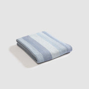 The Herringbone Hues Throw Blanket by In2Green, made in the USA from recycled cotton, boasts a cozy design with folded blue and white stripes on a white background.