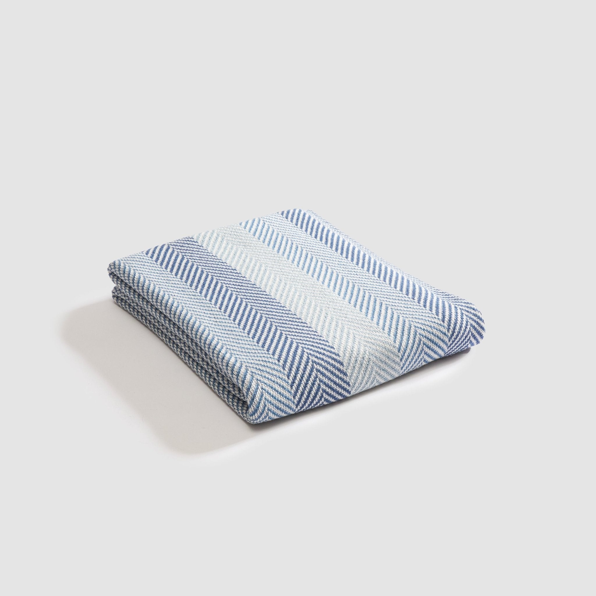 The Herringbone Hues Throw Blanket by In2Green, made in the USA from recycled cotton, boasts a cozy design with folded blue and white stripes on a white background.