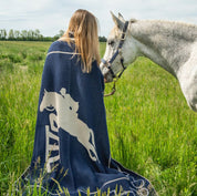A person with long hair sits in a grassy field facing a majestic white horse, wrapped in the in2green Equestrian Jumper Throw Blanket adorned with a horse pattern.