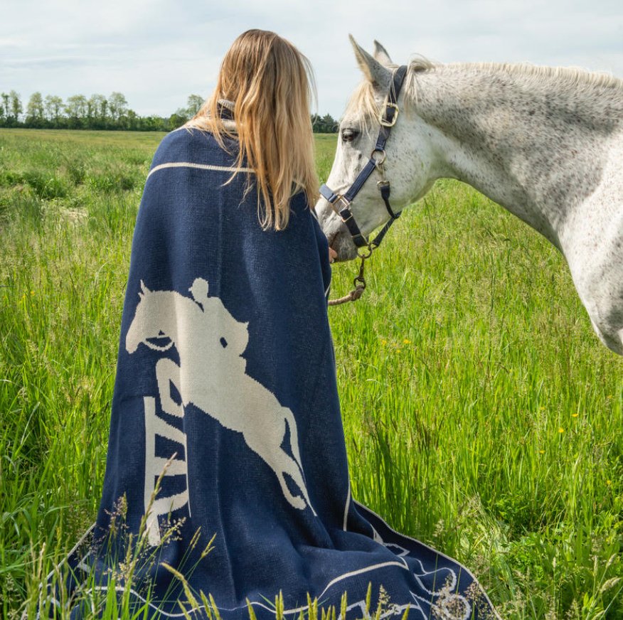 A person with long hair sits in a grassy field facing a majestic white horse, wrapped in the in2green Equestrian Jumper Throw Blanket adorned with a horse pattern.
