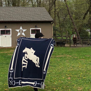 A person holds the in2green Equestrian Jumper Throw Blanket, navy blue with a white horse design, in front of a barn while a horse stands gracefully in the background.