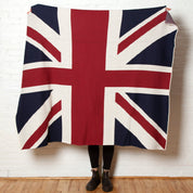 A person stands against a white brick wall, holding up the in2green Hometown Pride Flag - Cotton Sustainable Knit Blanket, featuring a Union Jack flag design and made in the USA from recycled cotton. Only their lower legs and feet are visible.