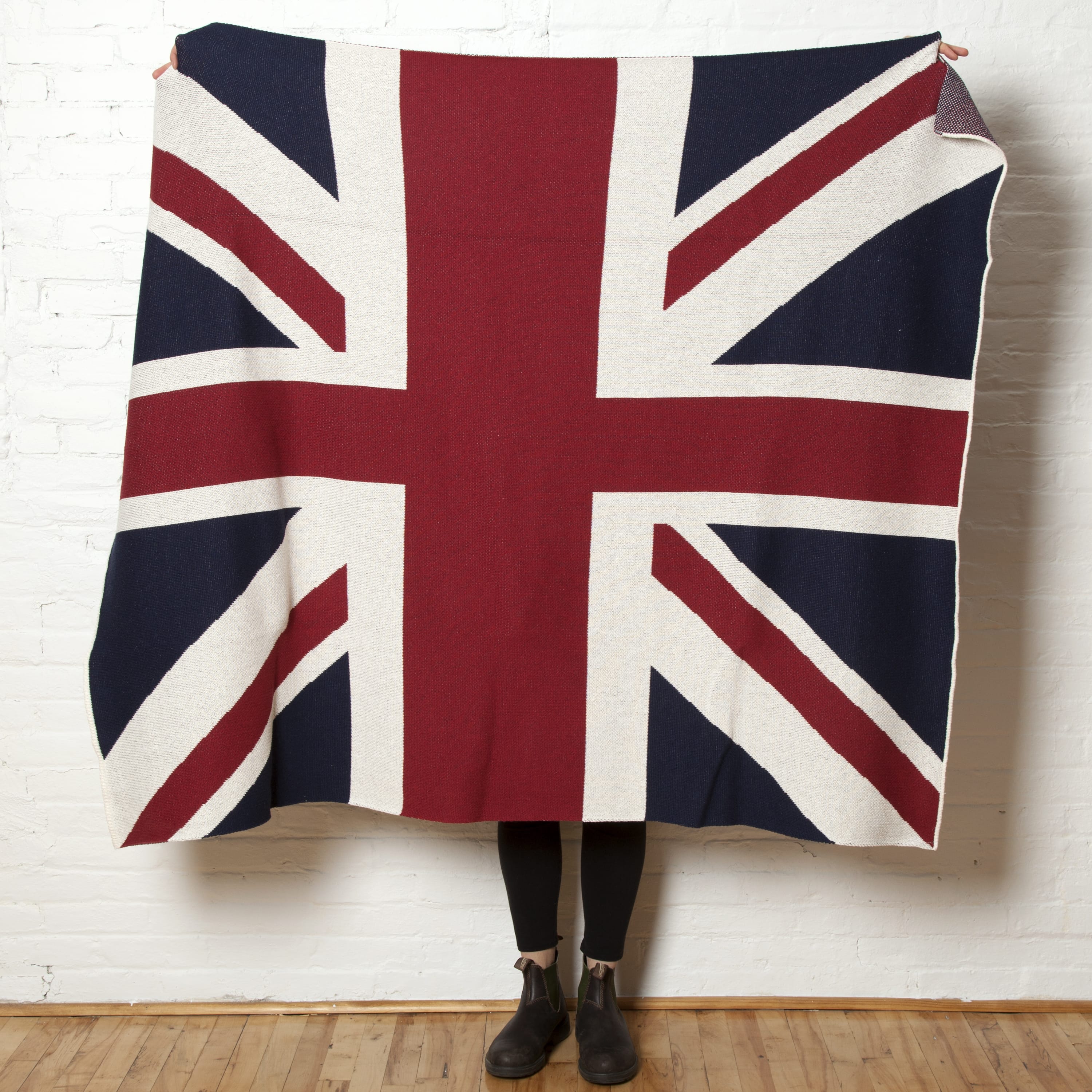 A person stands against a white brick wall, holding up the in2green Hometown Pride Flag - Cotton Sustainable Knit Blanket, featuring a Union Jack flag design and made in the USA from recycled cotton. Only their lower legs and feet are visible.