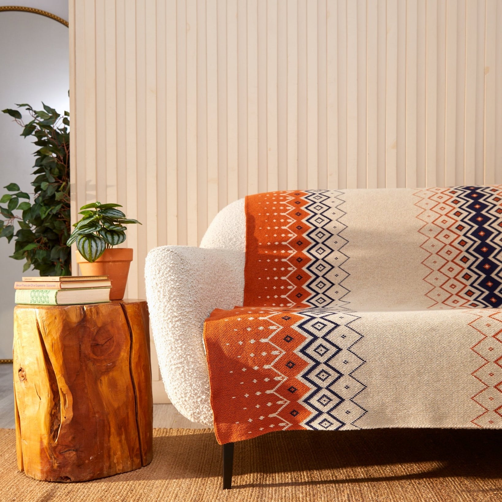 In a cozy living room, a white sofa is adorned with the in2green Vintage Icelandic Throw Blanket, crafted from recycled cotton and showcasing a charming orange and beige pattern. On a wooden side table rest books and a plant, while a tall plant and mirror complete the serene scene in the background.