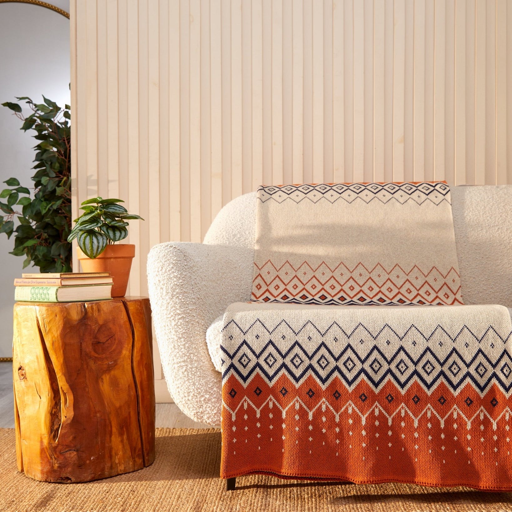 A cozy living room showcases a textured white sofa decorated with an in2green Vintage Icelandic Throw Blanket, enhancing the warmth and style. The space is charmingly completed with a wooden side table, books, a plant, and a round mirror.