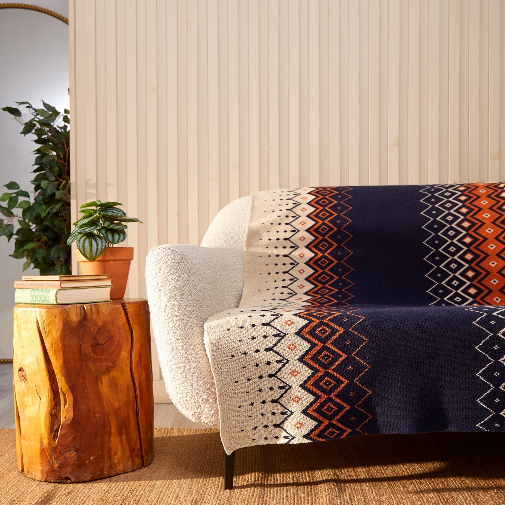 A cozy sofa adorned with an in2green Vintage Icelandic Throw Blanket crafted from recycled cotton is positioned beside a wooden side table holding a plant and books. In the background, a potted plant and mirror stand against a light wood-paneled wall.