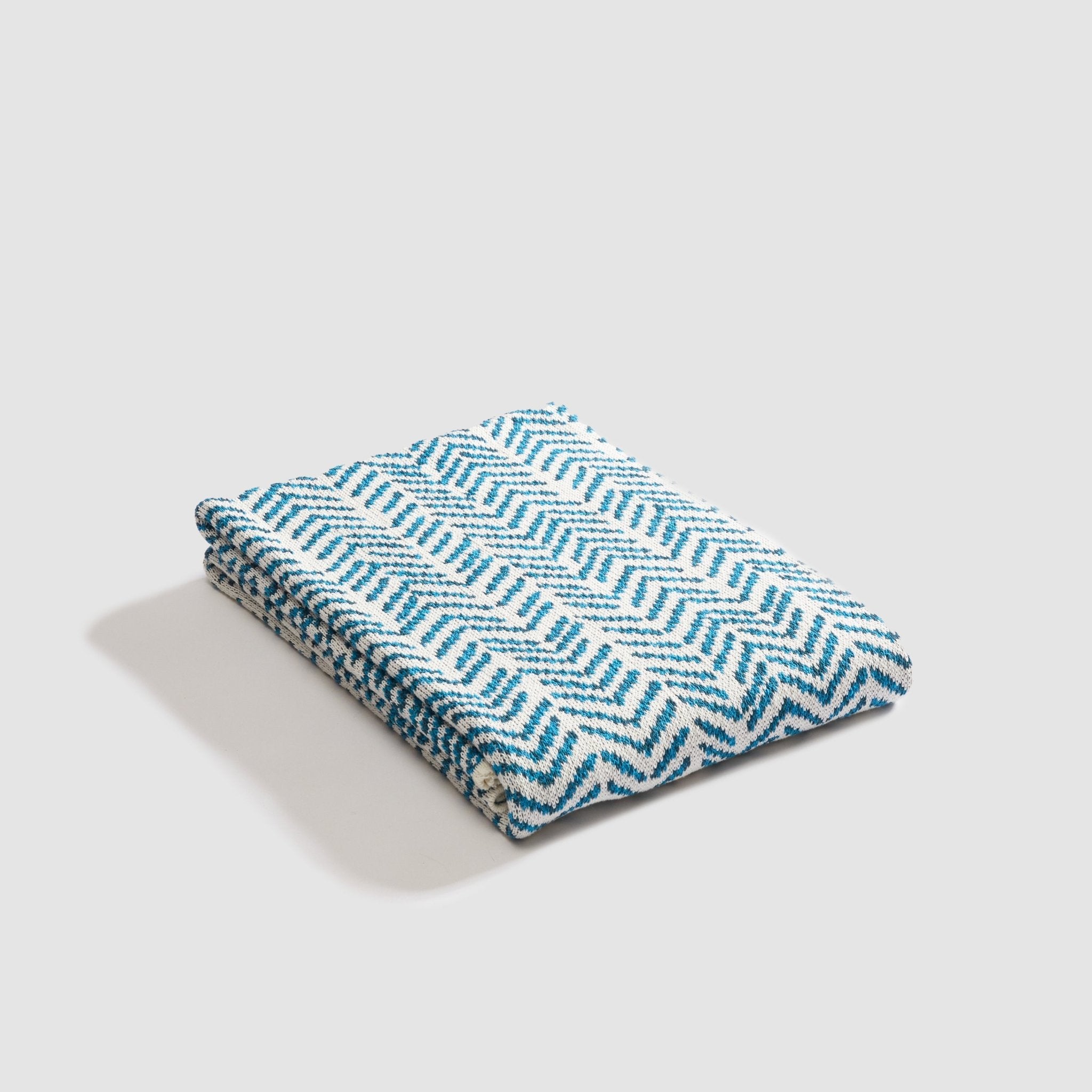 An In2Green Warrior Throw Blanket in teal, showcasing a bold blue and white zigzag pattern, lies folded against a plain backdrop, symbolizing strength and offering comfort.
